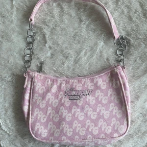 Rosa axelväska med Powerpuff Girls-mönster - Säljer en söt rosa axelväska med Powerpuff Girls-mönster. Väskan har en dragkedja upptill och en axelrem med kedjedetaljer. Perfekt för att ge en nostalgisk touch till din outfit!