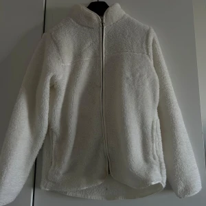 Vit teddyjacka med dragkedja - Mysig vit teddyjacka med dragkedja framtill. Perfekt för kyliga dagar med sin fluffiga och varma design. Jackan har långa ärmar och en hög krage för extra värme.