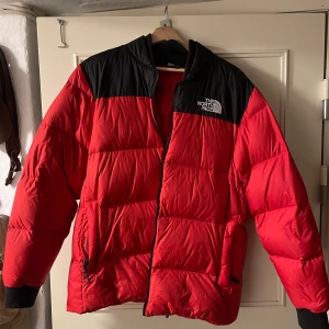 Röd dunjacka från The North Face - Säljer en röd och svart dunjacka från The North Face. Jackan har en broderad logga på bröstet och ryggen, samt en dragkedja framtill. Den är långärmad och har ribbade muddar. Perfekt för kalla dagar med sin 700-fill power.