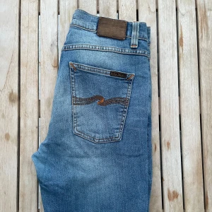 Blå jeans från Nudie Jeans - Snygga blå jeans från Nudie Jeans med klassisk femficksdesign och en cool broderad detalj på bakfickan. Perfekta för en avslappnad stil med en touch av trend. Midjan är normal och passformen är slim. De är en avklippt designe