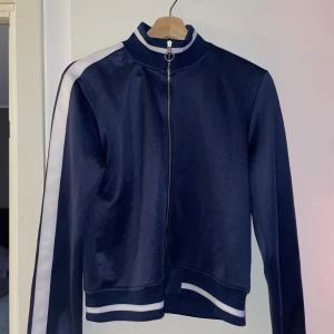 Mörkblå kofta  med vita ränder - Snygg mörkblå kofta med vita ränder längs ärmarna och dragkedja framtill. Jackan har en sportig look med ribbade muddar vid ärmslut och nederkant. Perfekt för en casual stil.