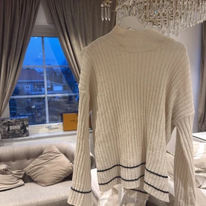 Vit ribbad stickad tröja - Säljer en stilren vit ribbad stickad tröja med svarta ränder vid ärmslut och nederkant. Tröjan har långa ärmar och en hög krage som ger en mysig känsla. Perfekt för kyliga dagar!