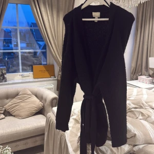 Svart kofta från H&M - Säljer en svart kofta från H&M med knytband i midjan. Perfekt för att hålla sig varm och stilren under kyligare dagar. Koftan har en enkel design med långa ärmar och är tillverkad i ett mjukt material.