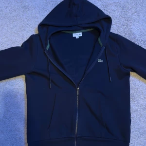 Mörkblå zip från Lacoste  - Säljer en stilren mörkblå hoodie från Lacoste med dragkedja och den klassiska krokodilloggan på bröstet. Tröjan har långa ärmar och en praktisk ficka framtill. Perfekt för en avslappnad look.