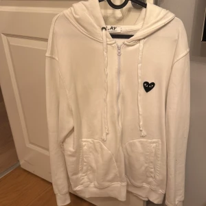Vit hoodie från Comme des Garçons Play - Säljer en stilren vit hoodie från Comme des Garçons Play med dragkedja och det ikoniska hjärtmärket på bröstet. Hoodien har en bekväm passform med långa ärmar och praktiska fickor framtill. Perfekt för en avslappnad look.