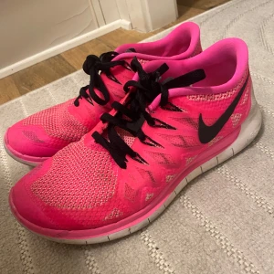 Rosa Nike sneakers - Snygga rosa Nike sneakers med svart snörning och vit sula. Skorna har en sportig design med meshdetaljer för extra andningsförmåga. Perfekta för träning eller en aktiv vardag.