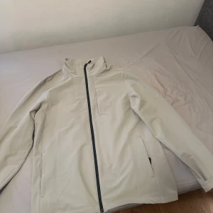 Beige vindjacka med dragkedja - Snygg beige vindjacka med hög krage och dragkedja framtill. Jackan har en minimalistisk design med två sidofickor och en bröstficka. Perfekt för blåsiga dagar.