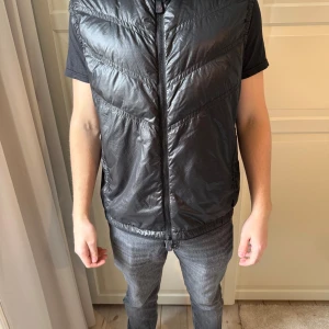 Svart dunväst från Moncler - Säljer en snygg svart dunväst från Moncler med dragkedja framtill. Västen har en stilren design och är perfekt för lager-på-lager under kyligare dagar. Den har en diskret logga på baksidan och är lätt att matcha med olika outfits. Storlek 4 men sitter bra på mig som bär storlek M i det mesta. Använd endast få tal gånger.