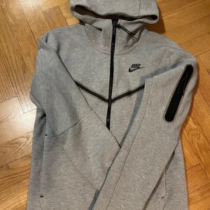 Grå hoodie från Nike - Säljer en stilren grå hoodie från Nike med dragkedja och huva. Tröjan har en svart Nike-logga på bröstet och en praktisk ficka på ärmen. Perfekt för en sportig look eller avslappnade dagar.