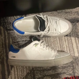 Vita sneakers från Axel Arigato - Snygga vita sneakers från Axel Arigato med blå och svarta detaljer. Skorna har en klassisk snörning och en diskret logga på sidan. Perfekta för en stilren look. Självklart äkta storlek 42 men passar 43. Pris ej hugget i sten vid snabbaffär 
