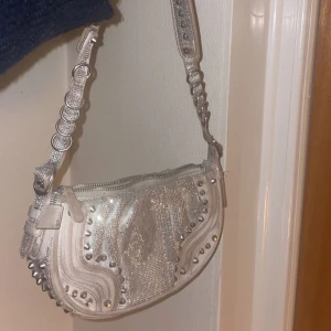 Silvrig axelväska med nitar  - Snygg silvrig axelväska med coola nitar och detaljer. Väskan har en unik design med en kedja som axelrem och en dragkedja för säker förvaring. Köpt på Zara i Paris. 299 kr + frakt 🫶 (går även hämta upp i Göteborg)