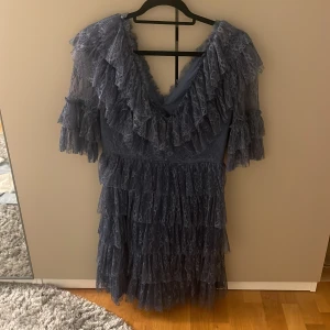 Indigoblå klänning från By Malina - Säljer en vacker indigoblå klänning från By Malina. Klänningen har volanger och spetsdetaljer med en v-ringad design. Perfekt för speciella tillfällen med sin eleganta look.