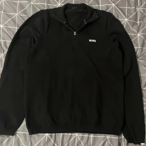 Svart tröja med dragkedja från Boss - Snygg svart tröja från Boss med halv dragkedja och broderad logga på bröstet. Perfekt för en stilren look. Tröjan har långa ärmar och en bekväm passform.