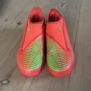 Adidas predator Röda och gröna fotbollsskor från Adidas - Snygga fotbollsskor från Adidas i en iögonfallande röd och grön färgkombination. Skorna har en strömlinjeformad design utan snörning för en tight passform. Perfekta för att dominera på planen med stil. Adidas predator. Färgerna är starkare neon än på bilderna för fick inte upp färgen på bilden så bra.