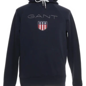 Mörkblå hoodie från GANT - Snygg mörkblå hoodie från GANT med logotyp och emblem på bröstet. Tröjan har en klassisk design med dragsko i huvan och en stor ficka framtill. Perfekt för en avslappnad stil.