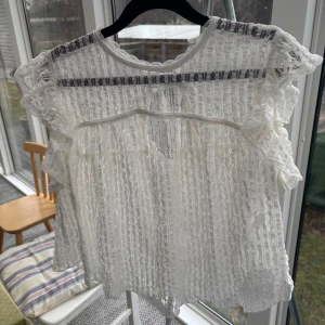 Vit spetsblus med volanger - Charmig vit spetsblus med volanger och knytning i nacken. Blusen har korta ärmar och ett luftigt, transparent mönster som ger en romantisk känsla. Perfekt för varma dagar eller som ett elegant lager över ett linne. 