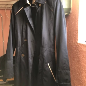 Mörkblå trenchcoat med bälte - Snygg mörkblå trenchcoat med knappar och bälte i midjan. Jackan har långa ärmar och stilrena detaljer i ljusare färg vid fickorna och längs ryggen. Perfekt för en elegant look.