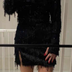 Svart glittrig klänning - Snygg svart klänning med glittriga detaljer och fransar. Perfekt för en kväll ute. Klänningen har en kort längd och långa ärmar, vilket ger en elegant look.