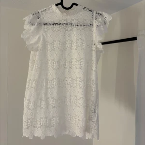 Vit spets topp - Charmig vit spets topp med korta ärmar och ett vackert blommigt mönster. Perfekt för somriga dagar eller speciella tillfällen. 