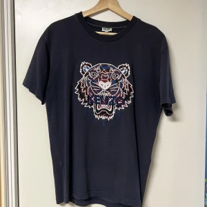 Mörkblå t-shirt med tigertryck från Kenzo - Mörkblå t-shirt från Kenzo med ett coolt tigertryck i färgerna blått, rött och vitt på framsidan. T-shirten har en normal passform och korta ärmar. Perfekt för en avslappnad stil.