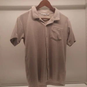 Beige kortärmad skjorta från Etab 1999 - Säljer en stilren beige kortärmad skjorta från Etab 1999. Skjortan har en klassisk krage och knappar framtill. Den är utrustad med en bröstficka för extra stil och funktionalitet. Perfekt för en avslappnad look.