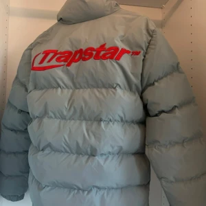 Grå pufferjacka från Trapstar - Säljer en grå pufferjacka från Trapstar med röd logga på bröstet och ryggen. Jackan har en dragkedja framtill och är tillverkad i 100% polyester. Perfekt för kyliga dagar med sin vadderade design.