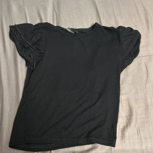 Svart topp från Boohoo - Säljer en svart topp från Boohoo med korta ärmar och volangdetaljer. Perfekt för en stilren look. Toppen är i ett mjukt material som känns skönt mot huden.