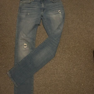 Blå jeans från Jack & Jones - Snygga blå jeans från Jack & Jones med slitna detaljer på låren och knäna. De har en klassisk femficksdesign och knappgylf. Perfekta för en avslappnad stil.