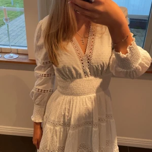 Vit klänning från By Malina - Säljer en vacker vit klänning från By Malina med spetsdetaljer och v-ringning. Klänningen har långa ärmar och en smickrande passform med smock i midjan. Perfekt för sommarens alla tillfällen!