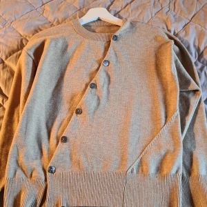 Beige kofta med knappar - Säljer en stilren beige kofta med unika diagonala knappar framtill. Tröjan har långa ärmar och ribbade muddar vid ärmslut och nederkant. Perfekt för en avslappnad och trendig look. I storlek L