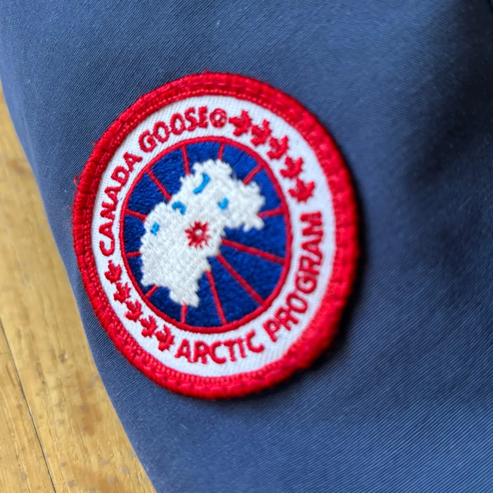 Säljer en blå vinterjacka från Canada Goose med huva och broderad logotyp på ärmen. Jackan har långa ärmar och flera praktiska fickor framtill. Perfekt för kalla dagar.. Takit.