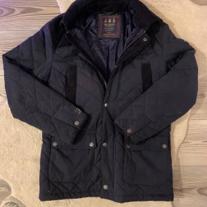 Navy quiltad jacka från Morris - Säljer en stilren svart quiltad jacka från Morris. Jackan har långa ärmar och en snygg broderad logga på axeln. Den är utrustad med både dragkedja och knappar framtill för extra stil och funktionalitet. Perfekt för lite uppklädda kyliga dagar.