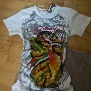 Vit t-shirt från Ed Hardy - Säljer en vit t-shirt från Ed Hardy med ett stort, färgglatt tryck av en kvinna och en drake. T-shirten har korta ärmar och är gjord i bomull. Perfekt för dig som gillar unika och iögonfallande designer!