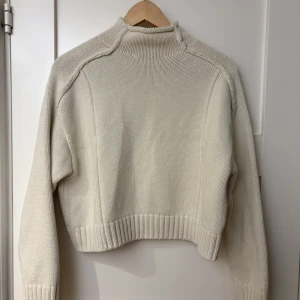 Vit stickad tröja från Zara - Säljer en stilren vit stickad tröja från Zara. Tröjan har en croppad design med långa ärmar och ribbade kanter. Perfekt för en avslappnad och trendig look. Passar bra till både jeans och kjol.