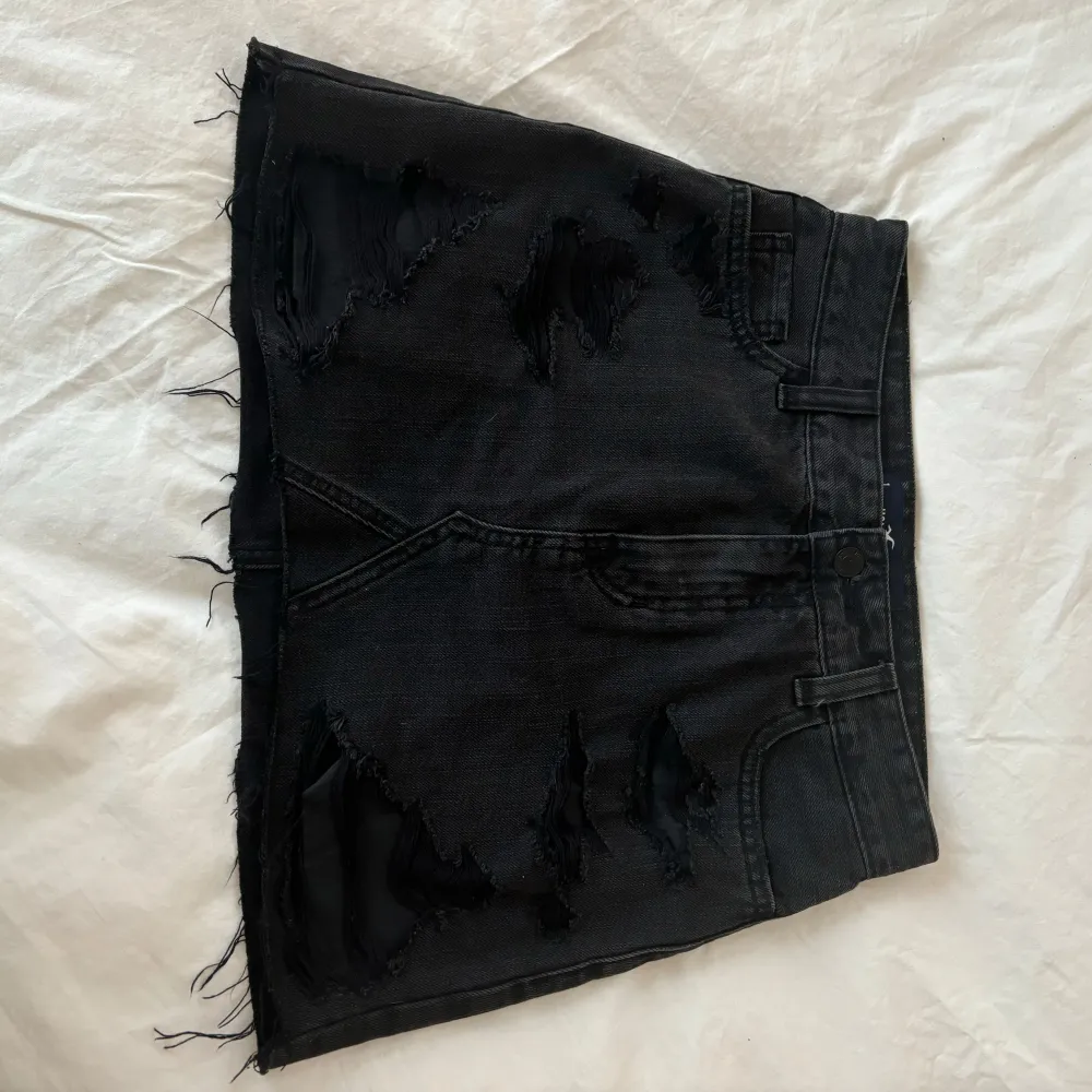 Snygg svart jeanskjol från Hollister med slitna detaljer och rå kant. Perfekt för en trendig look. Kjolen har en klassisk femficksdesign och är kort i modellen.. Hameet.