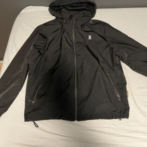 Svart jacka från Burberry - Säljer en stilren svart jacka från Burberry med dragkedja och huva. Jackan har en diskret logotyp på bröstet och är tillverkad i ett syntetmaterial. Perfekt för en elegant look.