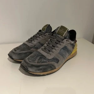 Valentino Rockrunner sneakers - Snygga Valentino Rockrunner sneakers i en mix av grått och svart med gula och röda detaljer. Skorna har en klassisk snörning och en stilren design som kombinerar olika material för en unik look. Priset är ej hugget i sten och är öppen för byte