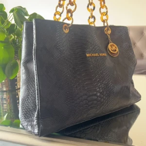Svart handväska från Michael Kors - Snygg svart handväska från Michael Kors med ormskinnsmönster och guldfärgade kedjehandtag. Väskan har en elegant design med en logotyp i guld på framsidan. Perfekt för att ge en lyxig touch till din outfit.