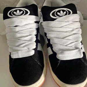 Svarta Adidas Campus sneakers - Snygga svarta Adidas Campus sneakers med vita detaljer och breda vita snören. Skorna har den klassiska tre-randiga designen på sidorna och en vit sula. Perfekta för en stilren och sportig look.