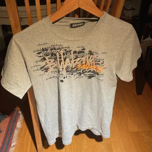 Grå t-shirt från Billabong - Säljer en grå t-shirt från Billabong med ett coolt tryck i svart och orange på framsidan. T-shirten är kortärmad och gjord av bomull, perfekt för en avslappnad stil. Passar bra till jeans eller shorts!