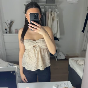 Beige axelbandslös topp med spetsdetaljer - Säljer en söt beige axelbandslös topp med spetsdetaljer och knytning framtill. Toppen har ett blommigt mönster och en elastisk överdel för bra passform. Perfekt för varma sommardagar!