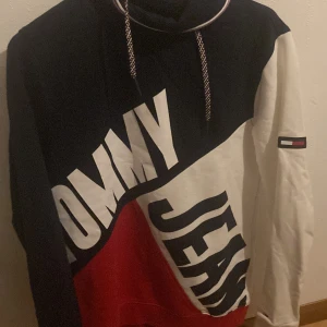 Flerfärgad hoodie från Tommy Jeans - Säljer en snygg flerfärgad hoodie från Tommy Jeans med stora bokstäver på framsidan. Tröjan har en kombination av svart, vit och röd färg och en praktisk huva med snörning. Perfekt för en avslappnad stil.