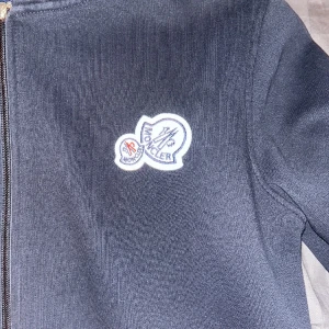 Svart huvtröja från Moncler - Säljer en stilren svart huvtröja från Moncler med dragkedja och broderad logga på bröstet. Tröjan har långa ärmar och praktiska fickor framtill. Perfekt för en casual look.