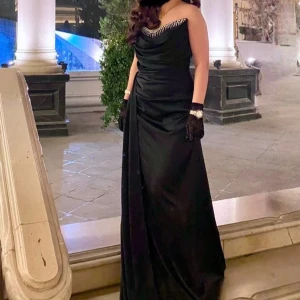 Svart långklänning med pärldetaljer - Elegant svart långklänning med pärldetaljer vid halsringningen. Klänningen har en draperad design och är ärmlös, vilket ger en sofistikerad look. Perfekt för en glamorös kväll.