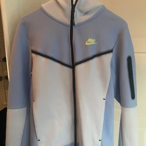 Ljusblå hoodie från Nike - Säljer en ljusblå hoodie från Nike med dragkedja och huva. Jackan har svarta detaljer och en gul Nike-logga på bröstet. Perfekt för en sportig look!