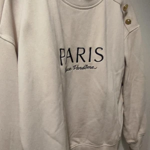 Beige sweatshirt från H&M - Säljer en beige sweatshirt från H&M med texten 'PARIS Place Vendôme' broderad på framsidan. Tröjan har långa ärmar och dekorativa guldknappar på axeln. Perfekt för en avslappnad stil.