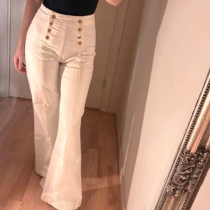 Vita högmidjade byxor med knappar - Snygga vita högmidjade byxor med guldknappar framtill. Byxorna har en bootcut-stil som ger en elegant siluett. Perfekta för att skapa en stilren look.