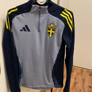 Sverige fotbolls tröja - Säljer en blå träningströja från Adidas med Sveriges landslagsemblem på bröstet. Tröjan har långa ärmar och en halv dragkedja framtill. Perfekt för fotbollsträning eller andra vardagliga aktiviteter