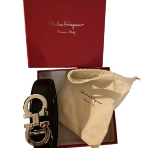 Svart ferregamo bälte  - Snyggt svart skärp med en stilren silverfärgad spänne i metall. Perfekt för att ge en elegant touch till din outfit. Passar bra till både jeans och kostymbyxor. (100-105 cm)