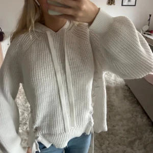 Vit stickad hoodie med snörning - Säljer en snygg vit stickad hoodie med snörning på sidorna. Tröjan har långa ärmar och en mysig huva med knytband. Perfekt för kyliga dagar och ger en stilren look. Passar bra till jeans eller leggings.